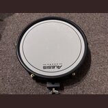 Kruhová elektronická bubenová pad v černé barvě s bílou úderovou plochou. Na spodní části plochy je černý nápis ALESIS DRUMS. Pad má černý obrubník a…