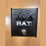 Černý kytarový efekt pedál s nápisem RAT v bílém rámečku. Horní část obsahuje tři černé otočné knoflíky s popisky DISTORTION, FILTER a VOLUME. V doln…
