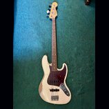 Bílá elektrická basová kytara s tmavě červeným pickguardem a hnedým hmatníkem. Na hlavě je vidět modré logo Fender a nápis Precision Bass. Povrch těl…