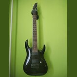 Černá elektrická kytara s tmavým hmatníkem, zavěšená na zelené stěně. Na hlavě nástroje je bílý nápis Ibanez. Kytara má dva humbuckery, jeden ovládac…