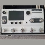 Bílý efektový procesor Hotone Ampero II Stage v hliníkovém kufru. Na horním panelu jsou nápisy AMPERO II STAGE, CDCM HD Gen 2 Tri-Core DSP Amp Modele…