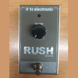 Stříbrný efekt pedál s nápisy RUSH a BOOSTER. Na pravé straně je logo tc electronic, vprostřed černý ovládací knoflík s popiskem VOLUME a červená kon…