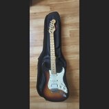 Elektrická kytara s tmelem v sunburst provedení a bílým pickguardem, položená na černém obalu. Na hlavě hmatníku je vidět logo a nápis Stagg. Nástroj…