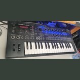 Černý syntetizátor s klaviaturou a ovládacím panelem s knoflíky a tlačítky. Na horní části je bílý nápis Roland JD-Xi a červeně svítící displej s tex…