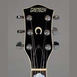 Detail hlavy elektrické kytary značky GRETSCH v černé barvě s bílým lemováním. Uprostřed hlavy je umístěno bílé písmeno C. Jsou vidět šest chromovaný…