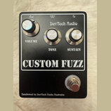 Černý efekt pedál s nápisem CUSTOM FUZZ a značkou DevTech Audio. Obsahuje tři stříbrné oválné knoflíky s popisky VOLUME, TONE a SUSTAIN. V horní část…