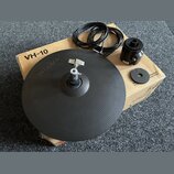 Černý gumový elektronický činel značky Roland, který leží na kartonovém obalu s nápisem VH-10. Součástí je černý kabel a černý plastový držák s nápis…