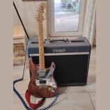 Elektrická kytara s hnědým dřevěným povrchem a bílým pickguardem stojí před černým kombem. Na hlavě kytary je nápis STRATOCASTER. Kombo má na přední…
