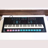 Černý elektronický klávesový nástroj s bílými a černými klávesami. Na horním panelu je nápis Music STATION, digitální displej zobrazující hodnoty 0.0…