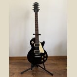 Černá elektrická kytara typu Les Paul značky Epiphone, umístěná na černém stojanu. Na hlavě hmatníku je bílý nápis Epiphone a vpravo pod ním kurzivní…