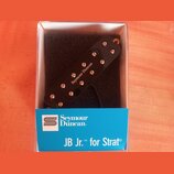 Černý humbucker snímač v plastovém průhledném boxu s modrou štítkem. Na krytce snímače je bílý nápis Seymour Duncan a na modrém štítku jsou loga a te…