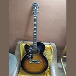 Akustická kytara s korpusem v tmavě hnědé a žluté barvě (sunburst). Na hlavě hmatníku je bílý nápis Epiphone. Kytara má ozdobné vkládky na hmatníku a…