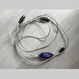 Stříbrný USB MIDI kabel s modrým plastovým převodníkem. Na převodníku je bílý nápis USB MIDI. Kabel má na koncích konektory USB a dva 5pinové DIN kon…