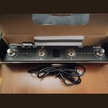 Černý elektronický přístroj s nápisy M-WAVE, HOST, USB a PEDAL. Na horní hraně jsou čtyři kovové konektory označené písmeny A, B, C a D, přičemž mezi…