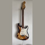 Elektrická kytara typu Stratocaster s černým lakem, který je na korpusu výrazně odloupaný a odhaluje světlé dřevo. Nástroj má bílý pickguard, tři sní…