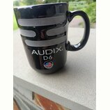 Černý keramický hrnek ve tvaru mikrofonu s vymalovanou stříbrnou mřížkou. Na povrchu je bílý nápis AUDIX. D6 a malá kulatá nálepka s vlajkou USA a ná…