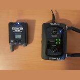 Na fotografii jsou dva černé přístroje značky LINE 6. Vlevo je vysílač s nápisy RELAY G50 TRANSMITTER a 2.4GHz DIGITAL WIRELESS SYSTEM, vybavený malý…