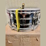 Stříbrný chromovaný buben s bílým koženem, který stojí na kartonovém krabici. Na přední straně je černé logo s nápisem „PD PERFORMANCE PERCUSSION“. K…