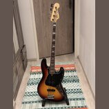 Elektrická basová kytara s korpusem v barevném přechodu od žluté přes oranžovou k černé (sunburst) a černým pickguardem. Na hlavě hmatníku je viditel…