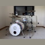 Bílá bicí souprava s modrým a červeným pruhem. Na přední straně basového bubnu je logo s nápisem SINCE 2004, DC DRUMS a QUALITY INSTRUMENTS. Souprava…
