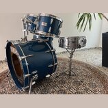 Modrá bicí souprava s kovovým hardwarem. Na basovém bubně je bílý nápis YAMAHA, na malém bubnu je viditelné logo YAMAHA. Na jednom z tomů je nalepený…