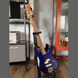Modrá elektrická basová kytara s bílým pickguardem, umístěná v černém stojanu. Na hlavě hmatníku je vidět nápis "Squier". Tělo nástroje má dva bílé s…