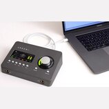 Šedý audio rozhraní s nápisem ARROW připojený bílým kabelem k notebooku MacBook Pro. Zařízení má digitální displej s logem UNIVERSAL AUDIO, ovládací…