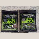 Dvě uzavřená balení kytarových strun v černých obalech s limegreenovými prvky. Na obou je nápis ERNIE BALL SLINKY COBALT ELECTRIC GUITAR STRINGS a te…