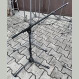 Fotografie zobrazuje několik černých kovových stativů na mikrofony s trojnožkou, umístěných na dlažbové ploše. Na jedné z nožek předního stativu je b…