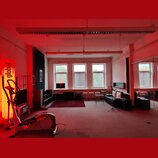 Fotografie zachycuje interiér místnosti s červeným osvětlením. V prostoru je umístěn černý digitální klavír s bílými klávesami a černá pohovka. V lev…
