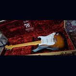 Elektrická kytara typu Stratocaster v barvě sunburst s bílým pickguardem a javorovým hmatníkem leží v otevřeném tvrdém kufru s červeným plyšovým vnit…