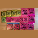 Sada různých balení kytarových a basových strun značky Ernie Ball. Horní řada obsahuje čtyři žluté balení s nápisem REGULAR Slinky a dvě růžové balen…