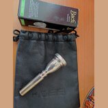 Stříbrný kovový moutur položený na černém koženém vakové pouzdře. Vedle nich je černá krabička s nápisy Bach Genuine Mouthpiece a LEAD FREE. Na moutu…