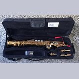 Zlatý saxofon v černém polstrovaném kufru. V horní části kufru je bílá nálepka s nápisem SAXOFON SERVIS JANA, QR kódem a kontaktními údaji. Vedle nás…
