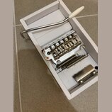 V bílém úzkém boxu je umístěn chromovaný kobylkový systém pro elektrickou kytaru s šesti sedly. Na každém sedle je vyryto slovo SADDLE a na základní…