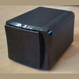 Černý, kompaktní reproduktor nebo subwoofer s hranatým tvarem a zaoblenými rohy. Na horní části je vyražený nápis "BASS" a na pravém boku je viditeln…