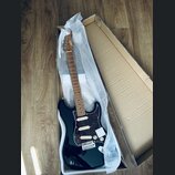Černá elektrická kytara s tmavě červeným šarlatanem (pickguard) v kartonové krabici s ochrannou bublinkovou fólií. Na hlavě hmatníku je bílý nápis "S…