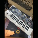 Bílo-šedý syntetizátor s klávesnicí a ovládacím panelem s mnoha posuvníky a tlačítky. Na horní části je nápis Roland GAIA SYNTHESIZER SH-01. Povrch p…