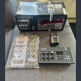 Na fotografii je elektronický modul Yamaha EAD10 v šedé barvě s ovládacími knofíky a displejem. Vedle něj je umístěn černý přívodní adaptér s nápisy…
