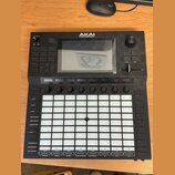 Černý MIDI kontrolér značky AKAI PROFESSIONAL s velkým displejem v horní části. Zařízení obsahuje mřížku 64 bílých podsvícených padů, několik ovládac…