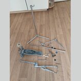Fotografie zobrazuje rozebraný chromovaný kovový stojan na hudební nástroj rozložený na dřevěné podlaze. Soubor obsahuje hlavní vrškový sloup s noham…