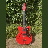Červená elektrická kytara s polodupatým korpusem a černým pickguardem. Na hlavě hmatníku je bílý nápis "Tonic". Nástroj má dva humbuckery, stříbrný m…