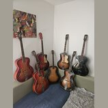 Skupina šesti archtop kytar v různých barvách a stavech, opřených o stěnu a pohovku. Nástroje mají dřevěné tělo s f-otvory, přičemž barvy sahají od č…