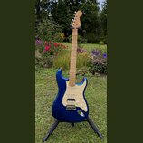 Modrá elektrická kytara typu Stratocaster s krčkem a hmatníkem ze světlého dřeva. Na hlavě je viditelné logo Fender. Kytara má krémový pickguard, tři…