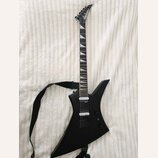 Černá elektrická kytara s ostře hranatým korpusem a černým popruhem. Na hlavě hmatníku je bílý nápis Jackson. Kytara má dva humbuckery, jeden ovládac…