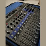 Šedý mixážní pult s modrými prvky a řadou posuvníků. Na panelu jsou viditelné nápisy jako GAIN, COMP, SEL, REC/PLAY a MUTE. Zařízení obsahuje početné…