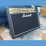 Kytarový kombo zesilovač značky Marshall v černém vinylovém potahu s bílým lemováním. Horní ovládací panel je zlatý a nese nápis JVM s řadou potencio…