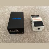 Bílo-černý ladicí pedál s nápisy OUTPUT, BYPASS, INPUT, Chromatic Tuner a TU-3. Na černé části je umístěno logo BOSS. Vedle pedálu je černá krabice s…