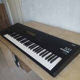 Černý digitální klavír s bílými a černými klávesami, umístěný na světlém dřevěném stole. Na pravé straně korpusu jsou bílé nápisy KORG a M1, pod kter…