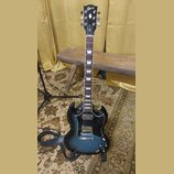 Černá elektrická kytara typu SG s modrým přechodem (sunburst) na korpusu. Na hlavě je bílý nápis Gibson a označení modelu SG. Nástroj má hnedý hmatní…